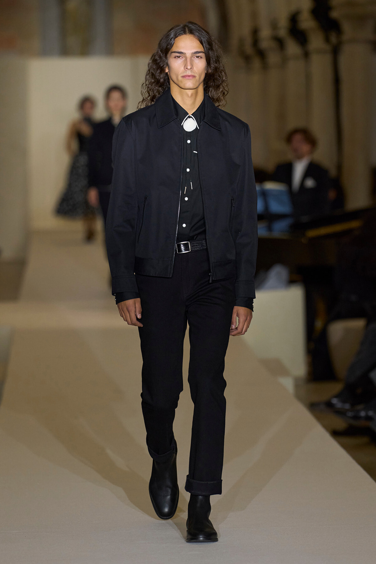 Male model walks the runway in an all-black outfit: black jacket, black shirt, black jeans, and black boots on a fashion show runway.
