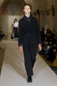 Male model in a black suit walks a fashion show runway with a polka-dot scarf.