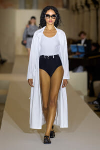 Model walks a runway wearing a white long coat over a white bodysuit and black high-waisted bottoms with a belt, accessorized with bold sunglasses and black flats.