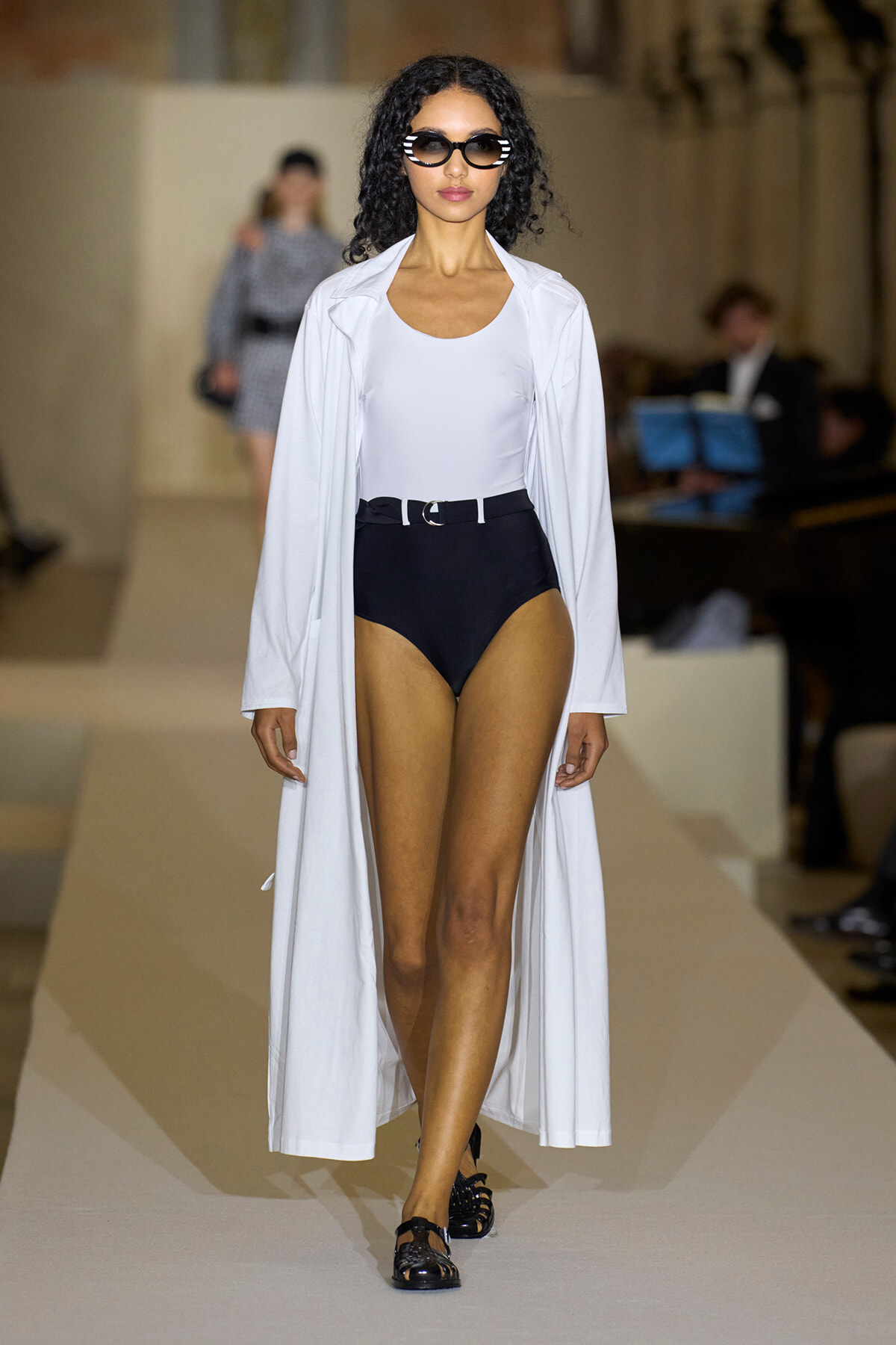 Model walks a runway wearing a white long coat over a white bodysuit and black high-waisted bottoms with a belt, accessorized with bold sunglasses and black flats.
