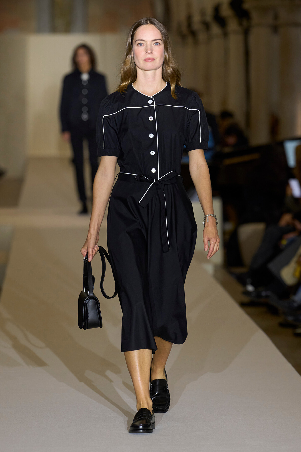 Model walks the runway in a black belted jumpsuit with white piping, carrying a small black bag.