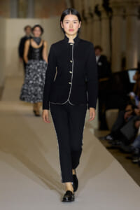 Model walking the runway in a black tailored suit with white piping and small gold buttons on the front.