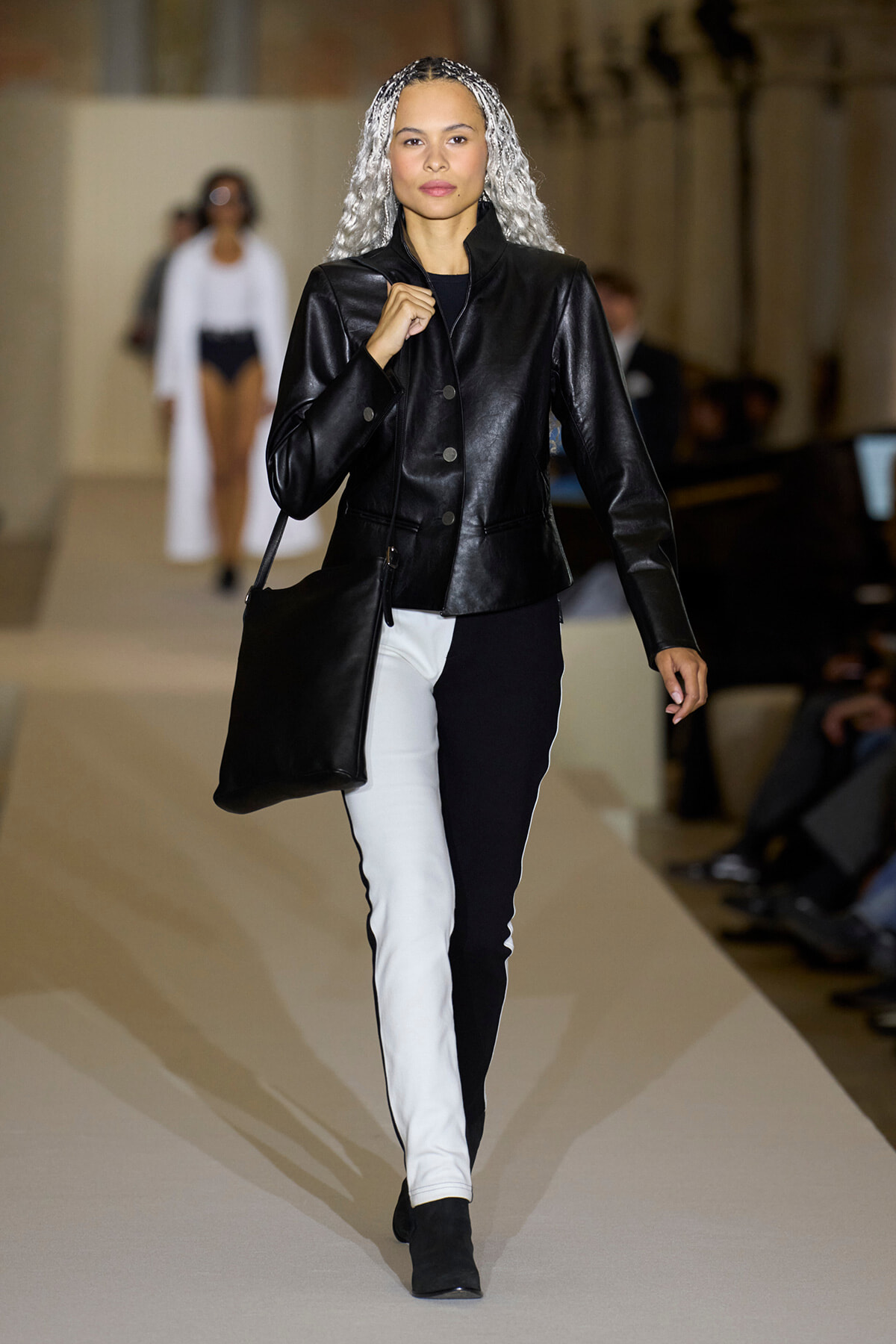 Model walking a runway in a black leather jacket, two-tone black-and-white pants, black ankle boots, and a large black shoulder bag, with silver braided hair.