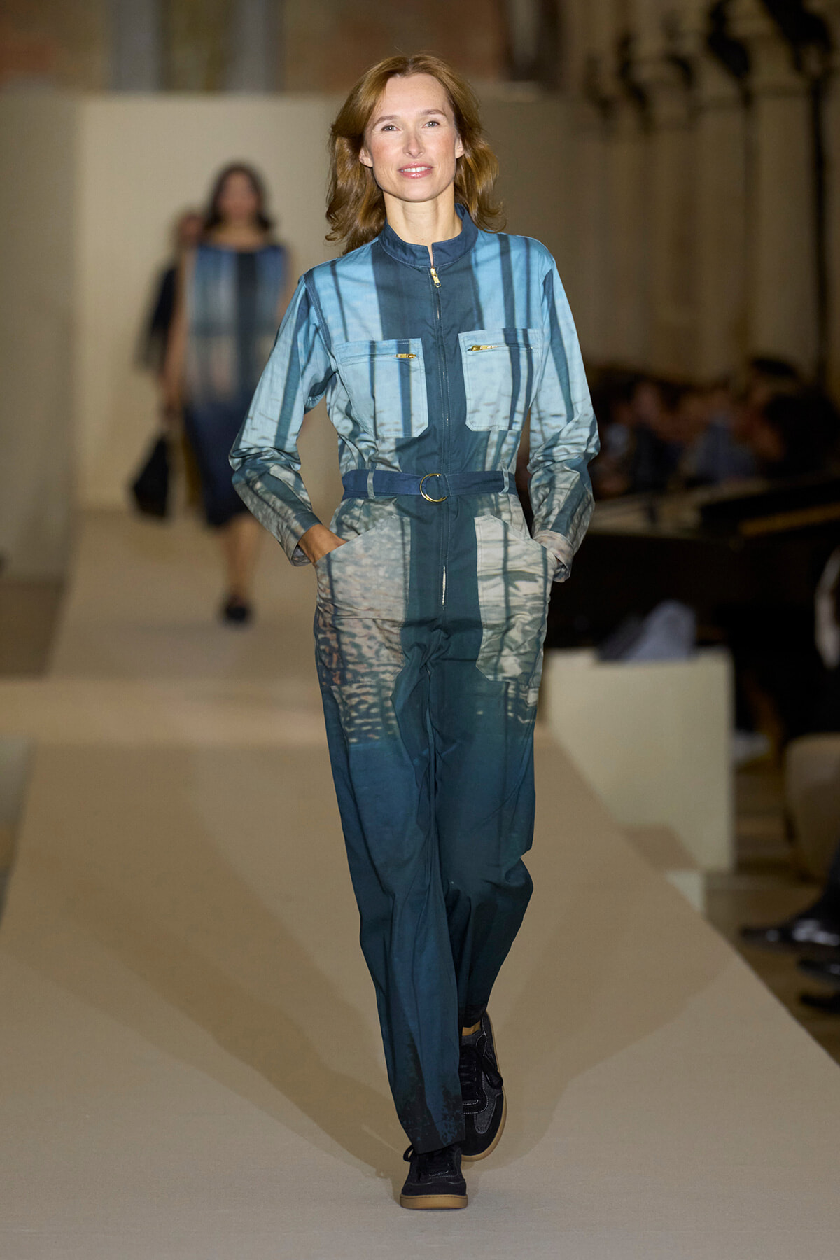 Female model walks the runway wearing a teal-blue, striped jumpsuit with a belt and front pockets.