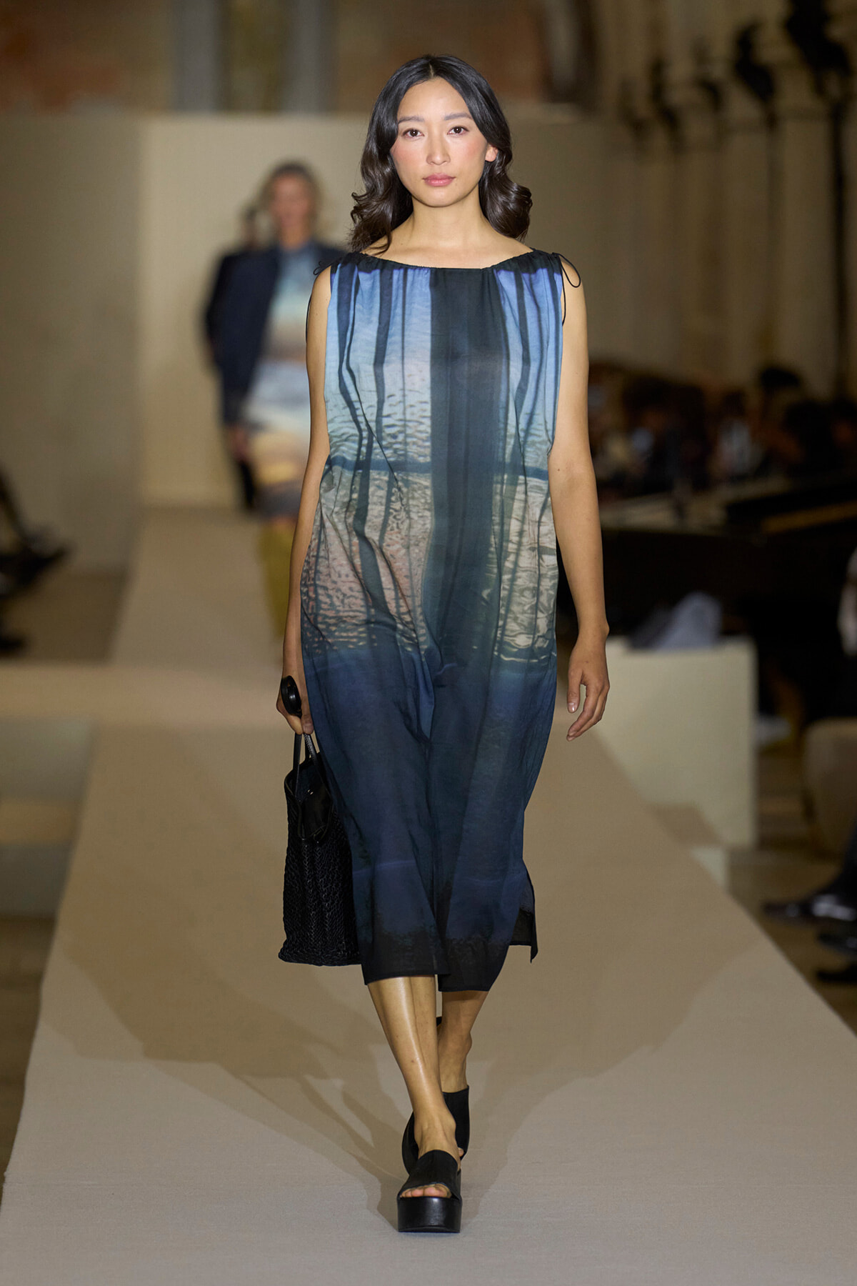 Model walks the runway in a sleeveless blue gradient dress, carrying a black handbag on a fashion show catwalk.