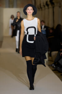 Runway model wearing a white sleeveless top with a bold black geometric print, a black mini skirt, knee-high socks, and a black hat, carrying a coat.