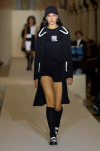 Model on a fashion runway wearing a black coat with white piping over a black top and shorts, a black cap, and knee-high socks.