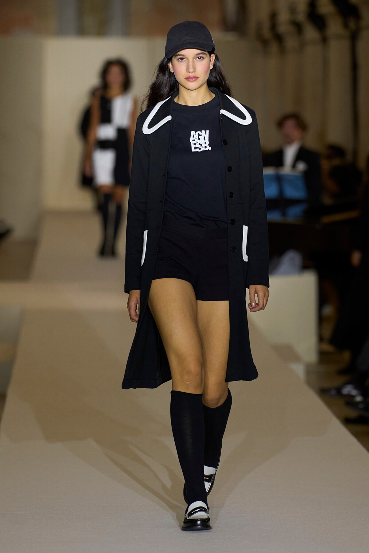 Model on a fashion runway wearing a black coat with white piping over a black top and shorts, a black cap, and knee-high socks.