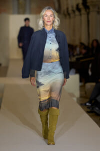 Model walking a fashion show runway in a gradient sunset-print midi dress with a navy jacket and mustard knee-high boots.