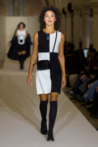 Model walks a runway in a sleeveless black-and-white color-block dress, black headband, knee-high socks, and a small black crossbody bag.