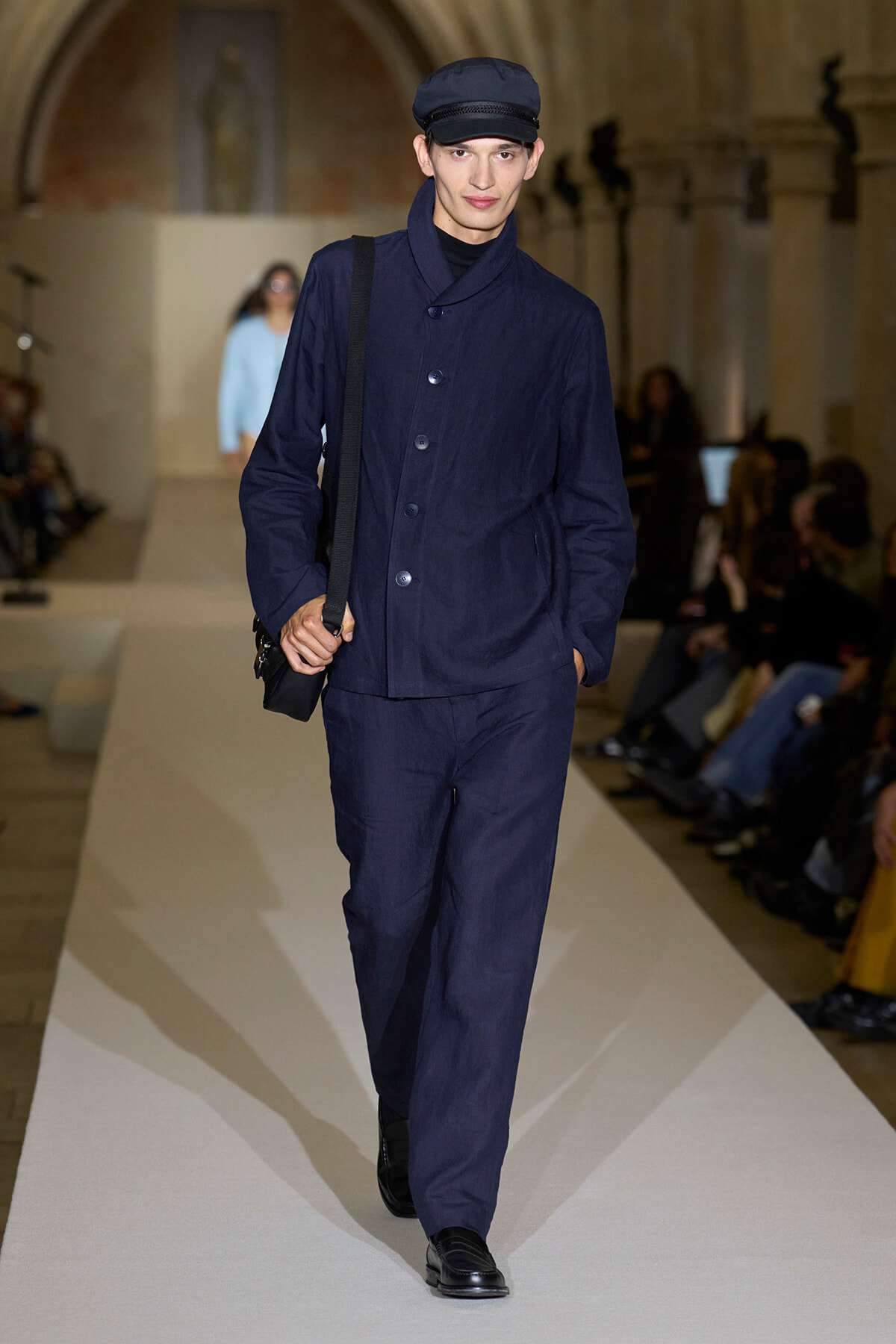 Male model on a runway wearing a navy blue coordinated outfit with a cap and black crossbody bag.