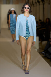 Model walking the runway in a light blue varsity-style jacket over a blue patterned swimsuit and white sunglasses.