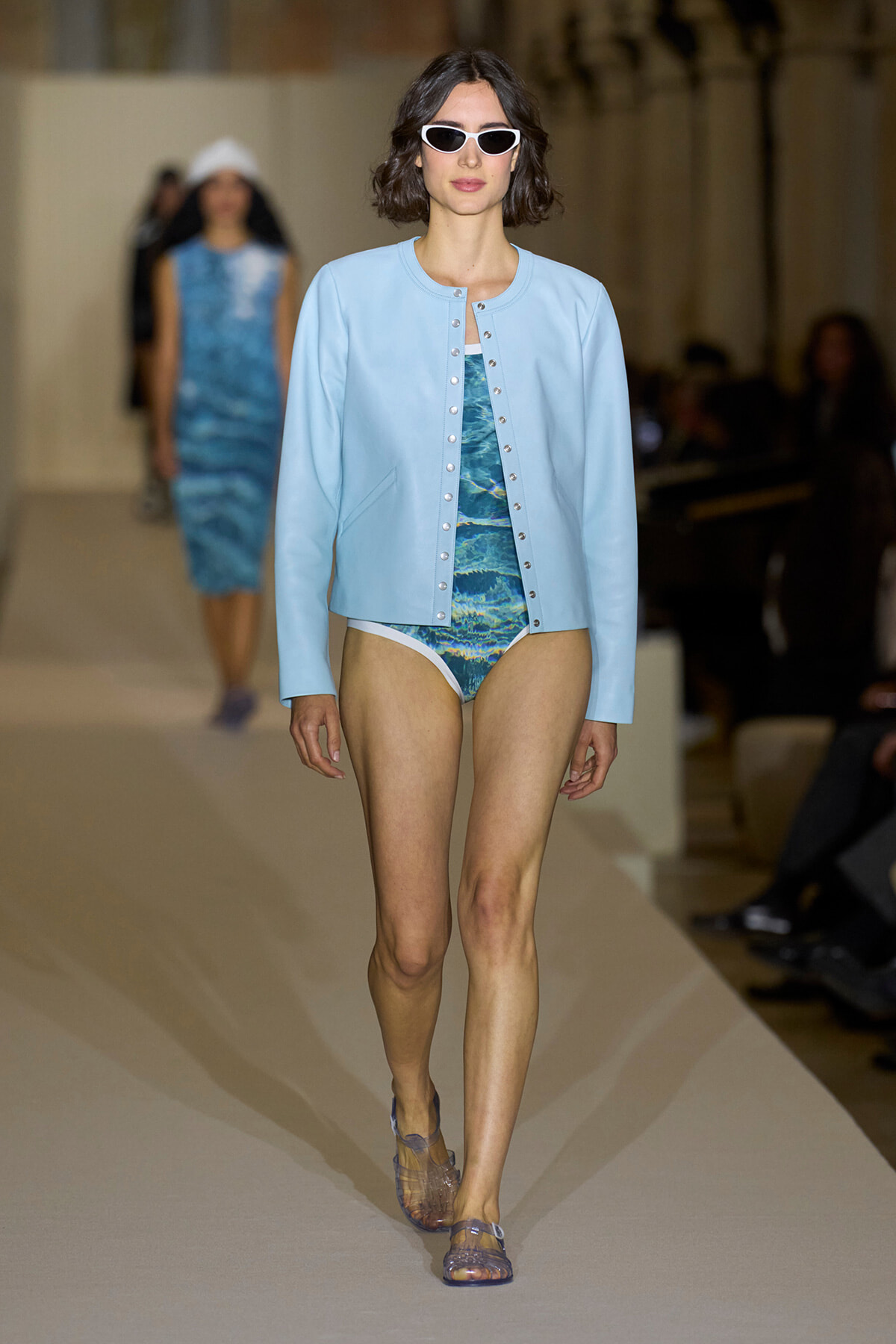 Model walking the runway in a light blue varsity-style jacket over a blue patterned swimsuit and white sunglasses.