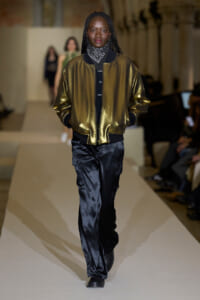 Model walks the runway in a shiny gold bomber jacket over a dark outfit, paired with black satin cargo pants and a patterned neck scarf.