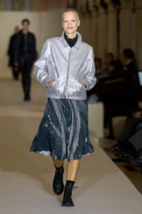 Model on a runway wearing a shiny silver bomber jacket, a dark patterned pleated skirt, and black combat boots.
