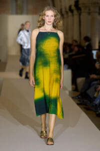 Model walks a runway wearing a sleeveless, green‑to‑yellow gradient midi dress with gold sandals and a pendant necklace, audience in background.