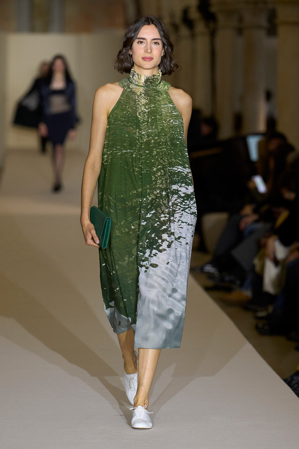 Model walking a runway in a sleeveless green gradient dress, holding a green clutch, wearing white sneakers.