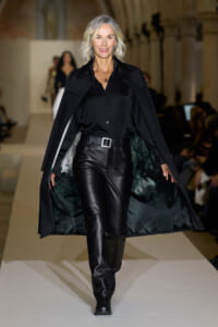 Model walks the runway in an all-black outfit: satin blouse, black leather pants, a wide belt, and a flowing cape with green-tinged lining.