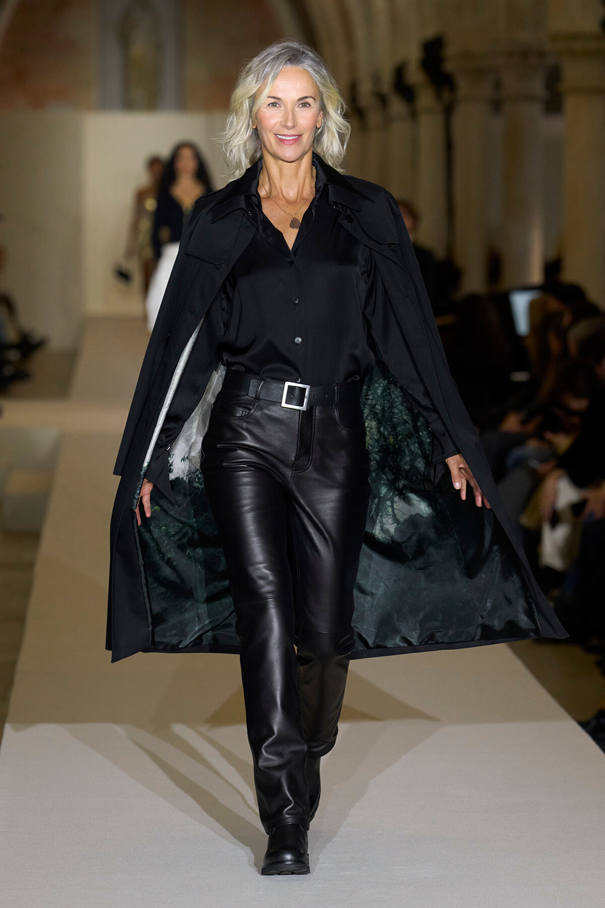 Model walks the runway in an all-black outfit: satin blouse, black leather pants, a wide belt, and a flowing cape with green-tinged lining.