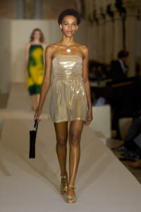 Female model on a fashion runway wearing a strapless metallic champagne mini dress, gold heels, and holding a black handbag.