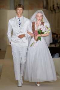 Bride and groom walking arm in arm on a runway; groom in white suit, bride in white gown with veil holding a pink bouquet of lilies.