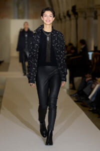 Model walking the runway in a black ensemble: mesh-knit top, jacket with a safety-pin print, black leather pants, and black ankle boots, wearing layered necklaces.