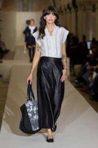 Model walking a runway in a white short-sleeve blouse and black leather midi skirt, carrying a two-tone gray and black tote bag in hand.