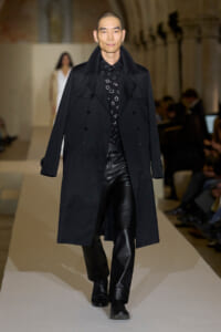 Male model on a runway wearing a long black trench coat over a black patterned shirt and black leather pants.