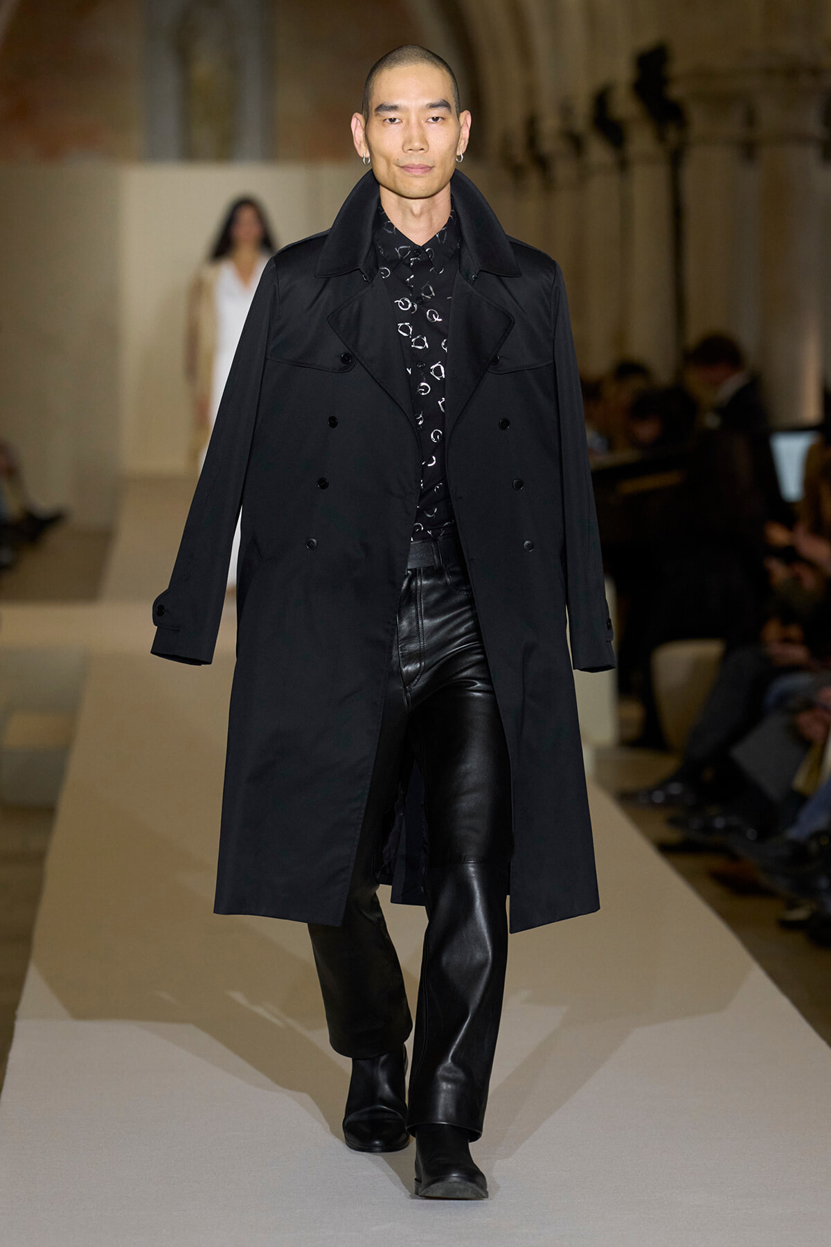 Male model on a runway wearing a long black trench coat over a black patterned shirt and black leather pants.