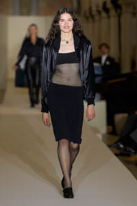 Runway model walking in a black sheer top under a satin bomber jacket, paired with a black midi skirt, fishnet tights, and black heels.