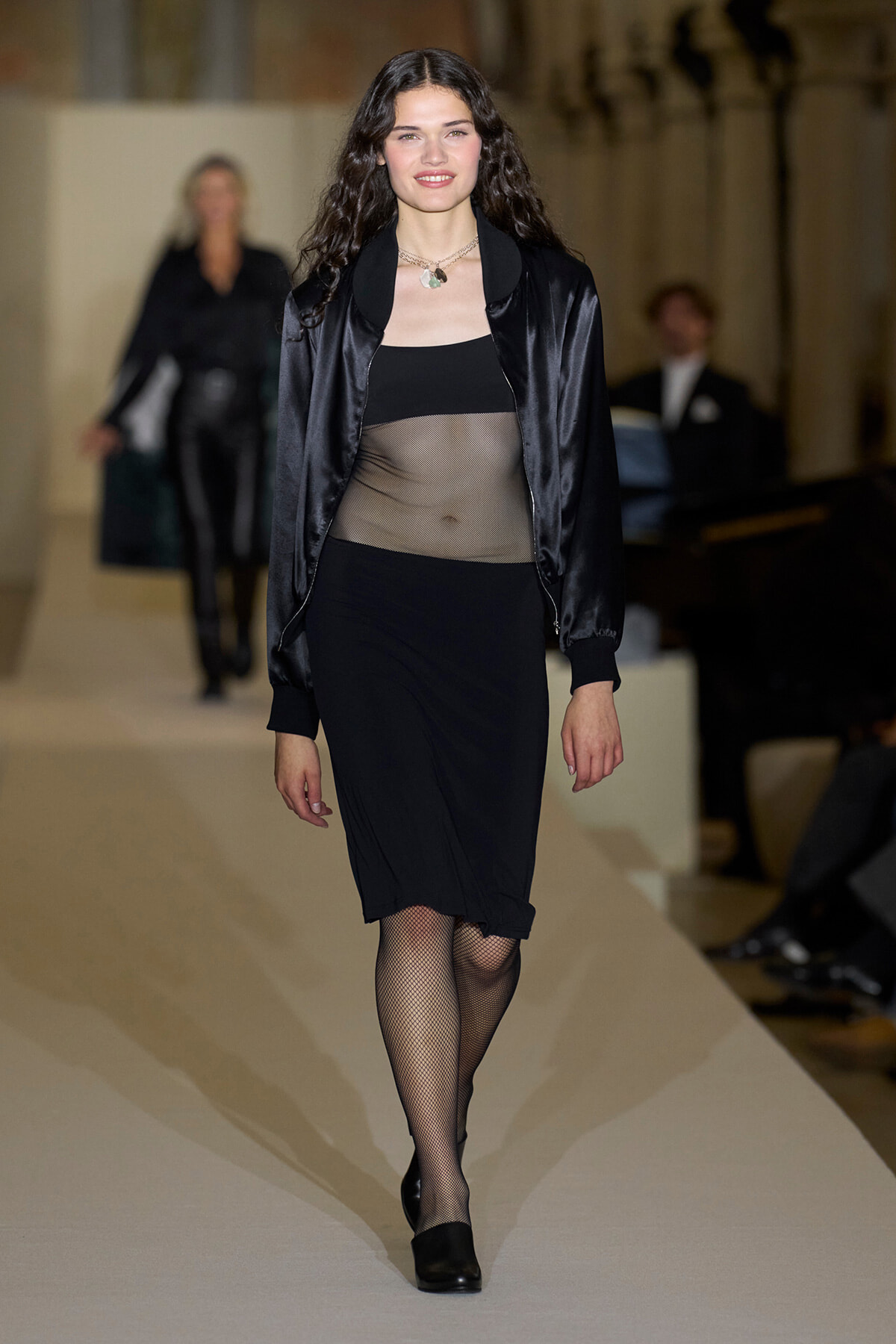 Runway model walking in a black sheer top under a satin bomber jacket, paired with a black midi skirt, fishnet tights, and black heels.