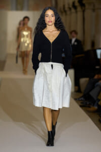 Model walks the fashion runway in a black zip-front V-neck top paired with a white textured knee-length skirt and black ankle boots, hands in pockets.