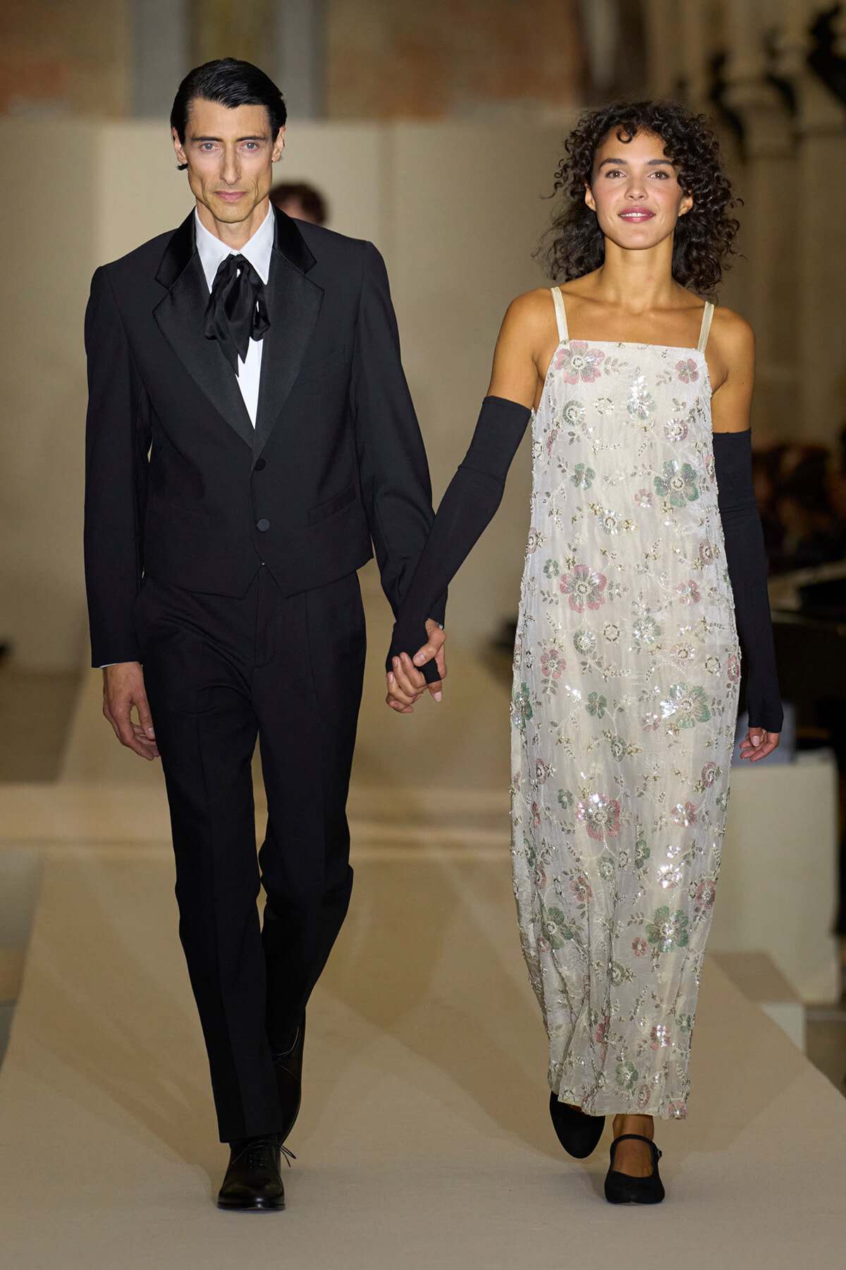 Two models hold hands and walk the runway: a man in a black suit with a bow tie and a woman in a pale floral sequin dress with black gloves.