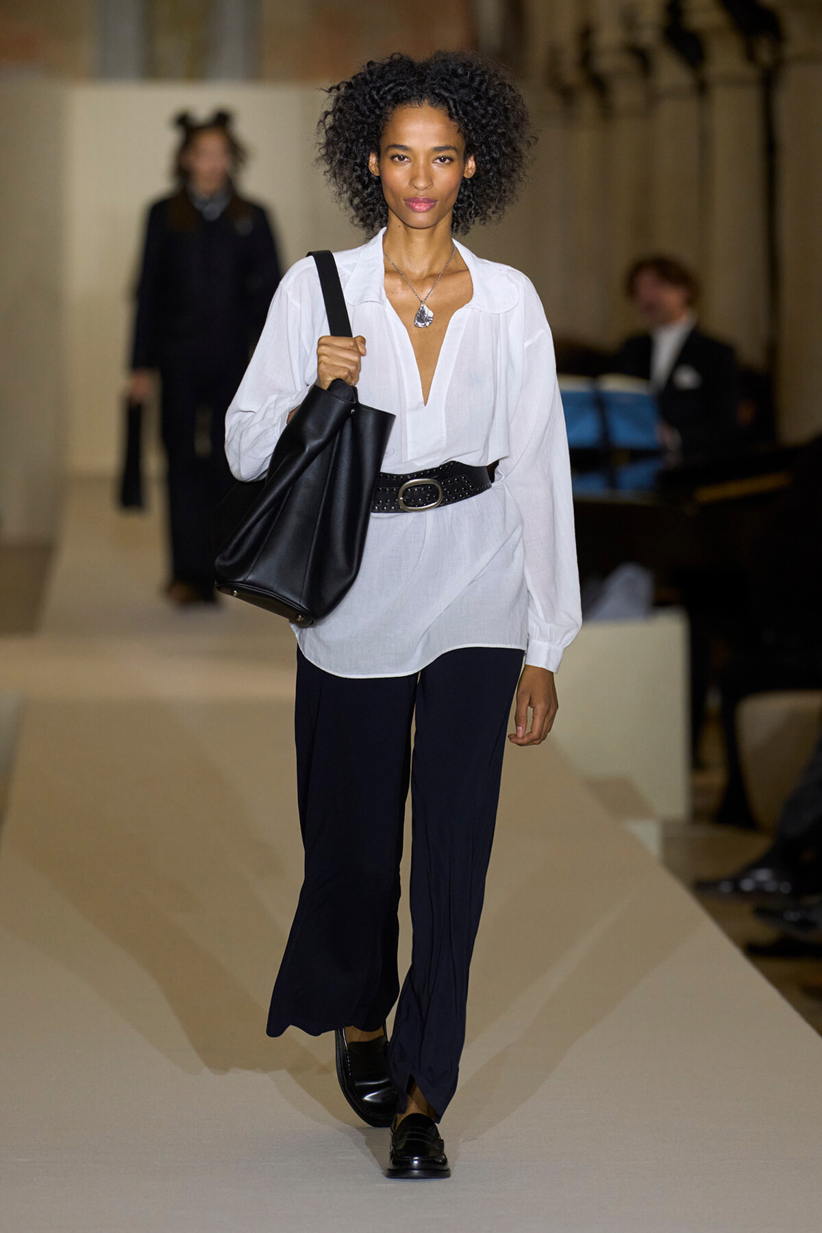 Model walking a runway in a white blouse with a deep V, cinched by a wide black belt, navy wide-leg pants, black loafers, and a large black tote over the shoulder.