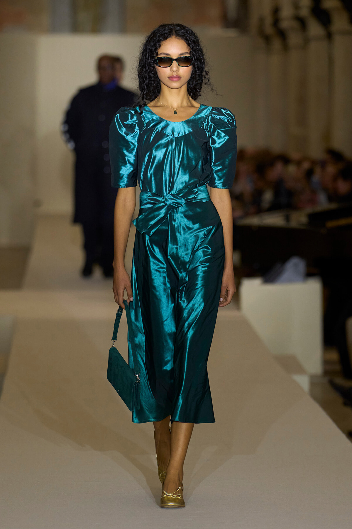 Model on a fashion runway wearing teal satin dress with puff sleeves and a waist bow, carrying a teal clutch and wearing sunglasses with gold heels.