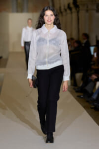 Model walks the runway wearing a sheer white zip-front blouse with black pants and a small black clutch with a gold chain.
