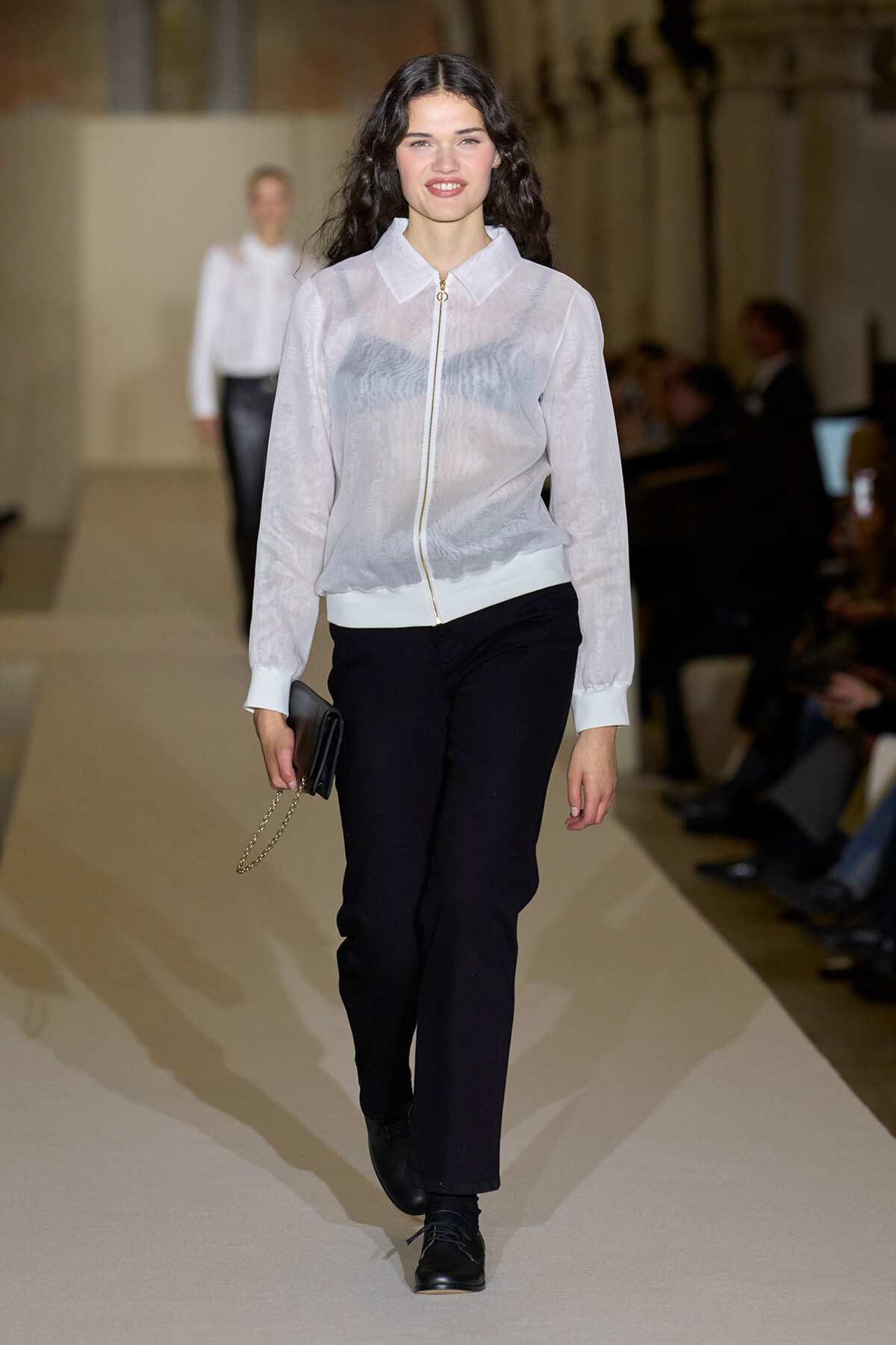 Model walks the runway wearing a sheer white zip-front blouse with black pants and a small black clutch with a gold chain.