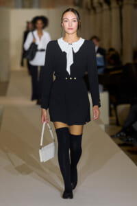 Model walking a runway in a black long-sleeve dress with a white bow-tied collar, gold belt accents, black knee-high socks, and a white handbag.