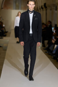 Male model walking a fashion runway in a black suit, white shirt, and black scarf accessory, with blazer pocket square visible.