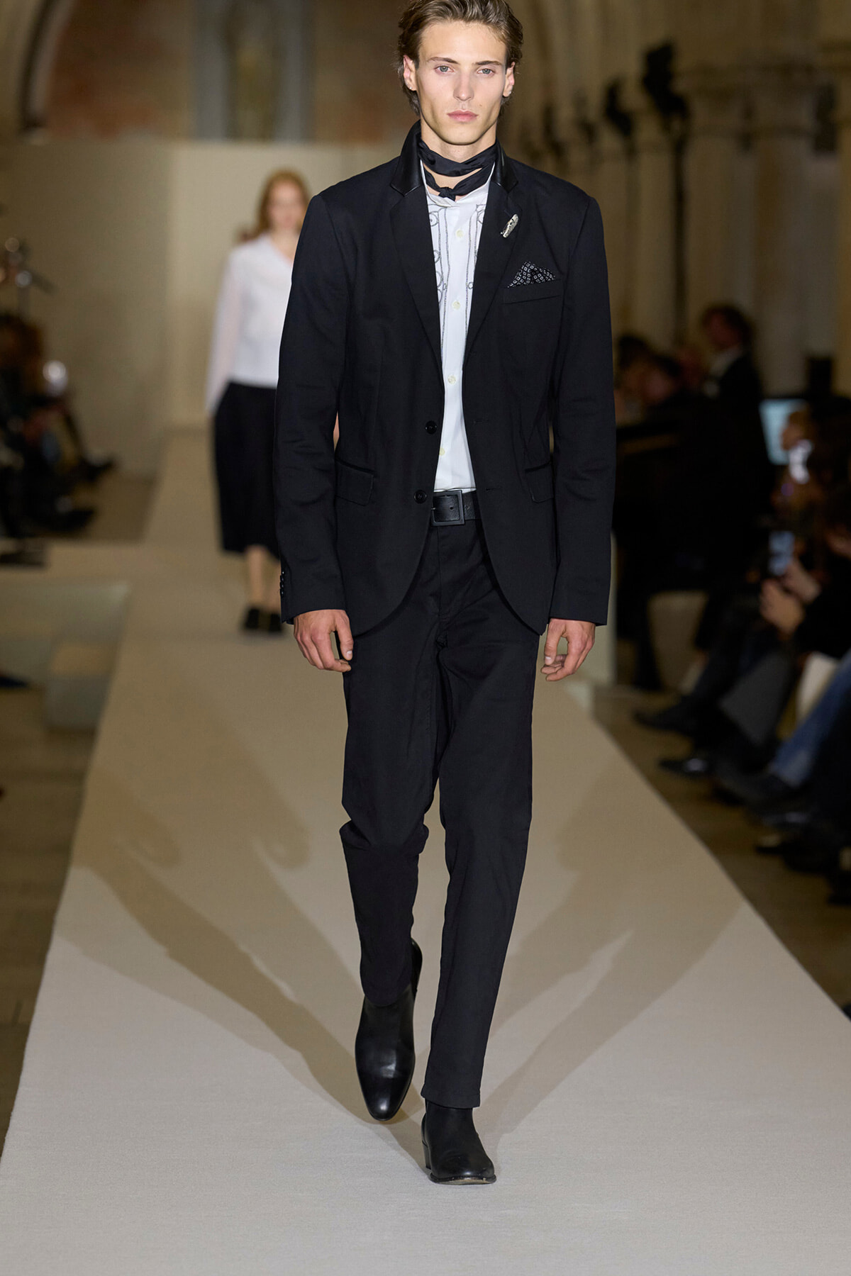 Male model walking a fashion runway in a black suit, white shirt, and black scarf accessory, with blazer pocket square visible.