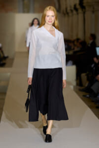 Runway model with long red hair wearing a white wrap blouse and black wide-leg skirt, carrying a black handbag.