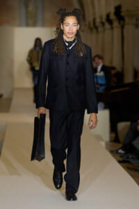 Male model walks the runway in a black utility-style suit, holding a black portfolio bag.