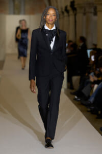 Model walks the runway in a tailored black suit with a white shirt and large black bow tie, hands in pockets.
