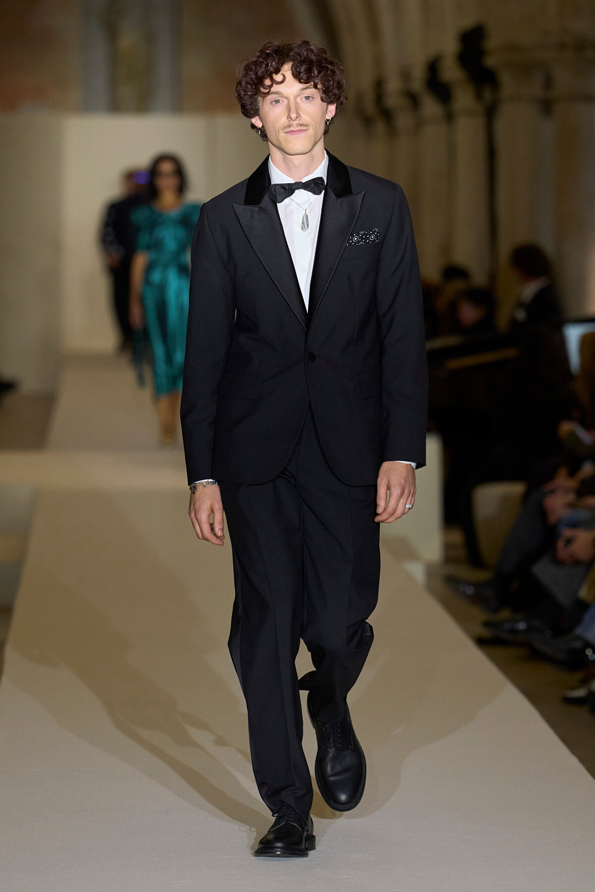 Male model walks the runway in a black tuxedo with satin lapels, white shirt, black bow tie, and pocket square.