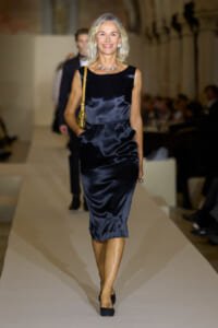 Model walking a fashion runway in a navy satin knee-length dress, with pearl jewelry and a gold-yellow shoulder bag.