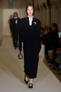 Model walks a runway in a black tailored suit with a white button‑up collar, gold brooch, black gloves, and a small black handbag; classic, minimalist ensemble.