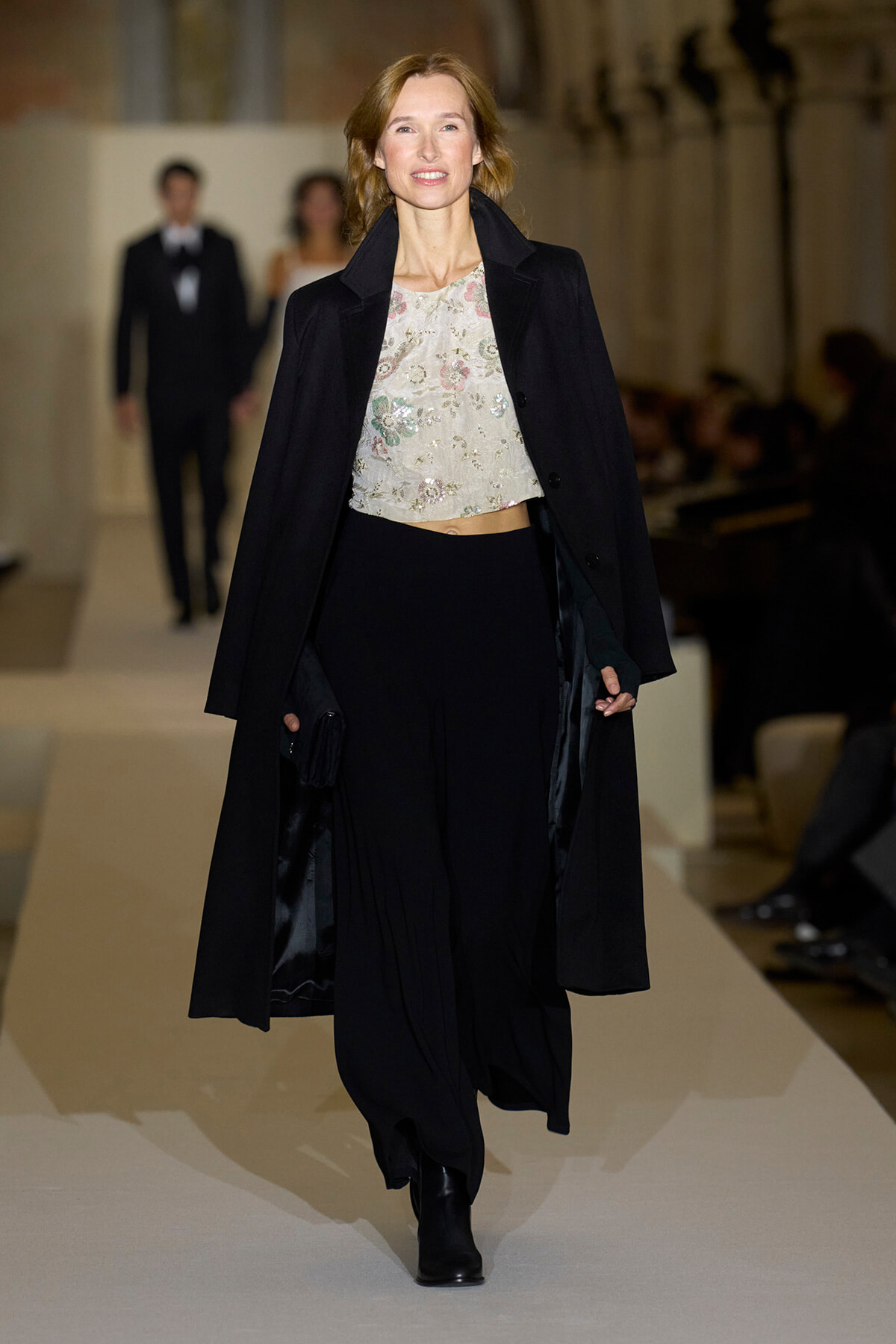 Full-length runway shot of a model wearing a long black coat over a pale floral crop top and black wide-leg pants, walking the catwalk.