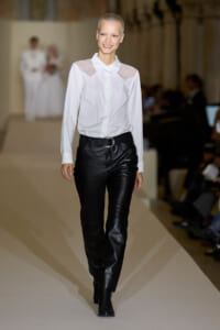 Model walks runway in a white button-up blouse with sheer shoulder panels and black leather pants with a belt.