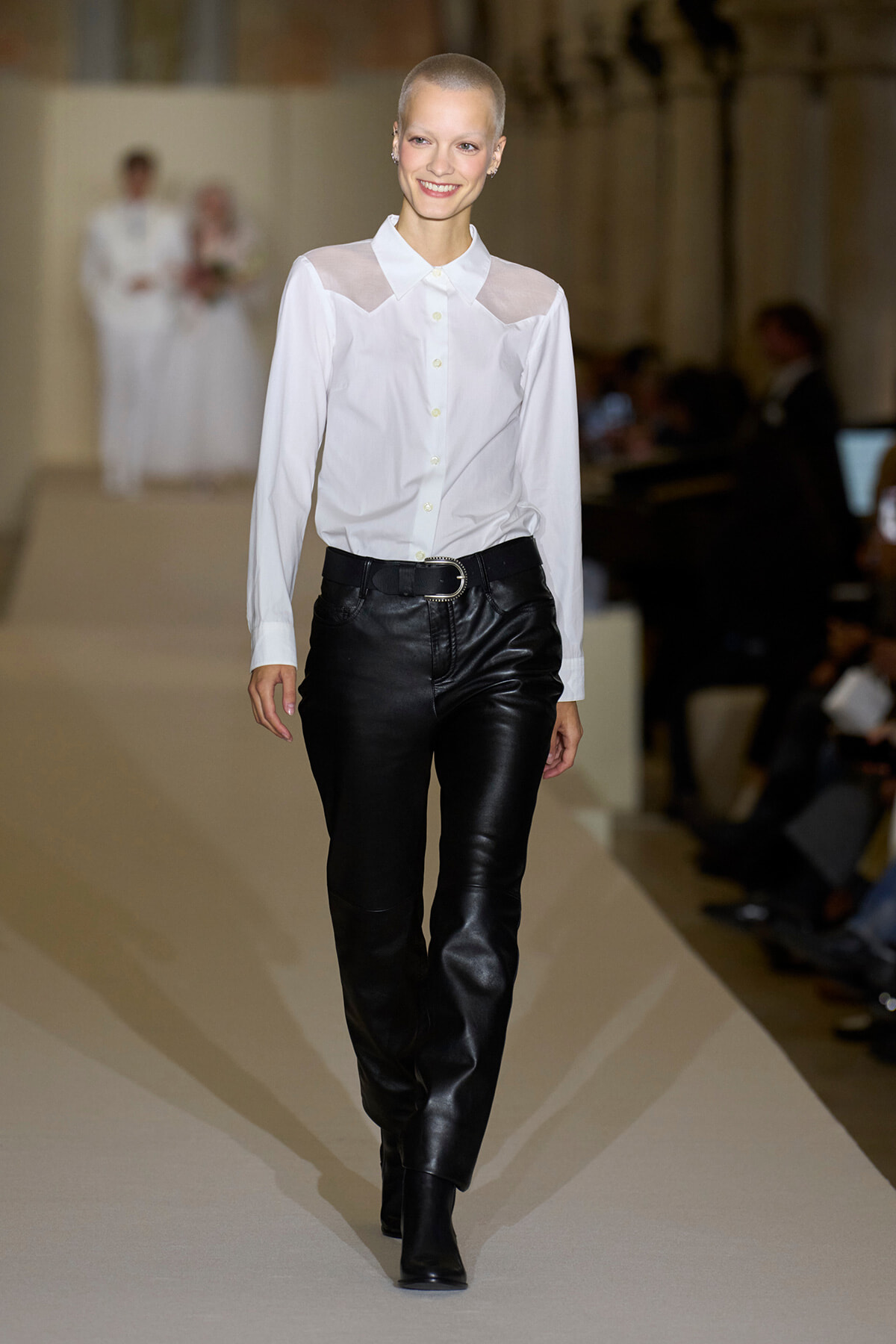 Model walks runway in a white button-up blouse with sheer shoulder panels and black leather pants with a belt.