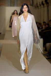 Model walks the runway in a white, V-neck satin gown with a thigh-high slit, draped beige shawl, and gold sandals.
